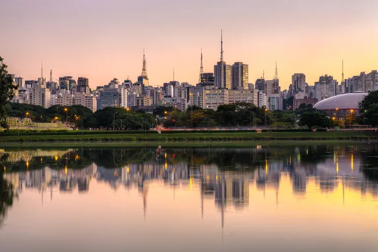 Skyline de São Paulo refletido no lago do Parque Ibirapuera ao entardecer, com a Oca à direita